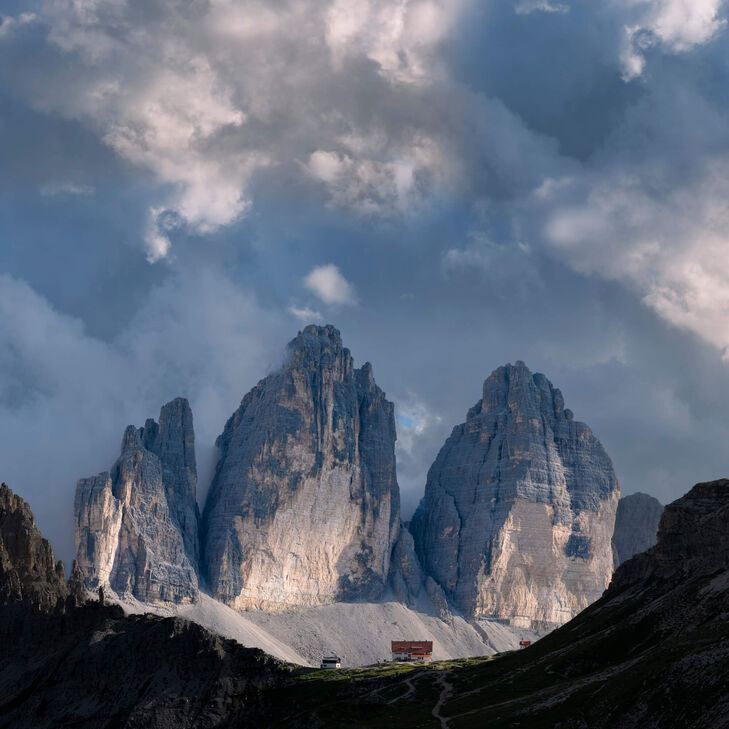 Ungewöhnliche Perspektive auf die kleine Drei-Zinnen-Hütte vor den riesigen Felsen der drei Zinnen. Dramatische Wolkenstimmung.