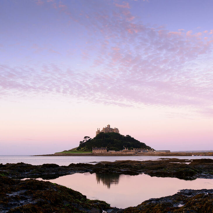 Der St. Michael´s Mount unter rosafarbenem Himmel bei Sonnenaufgang. Mit Spiegelung im Wasser zwischen den Steinen.