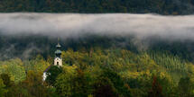 Sonnenbestrahlte Kirche von Ruhpolding vor der dunkelgrünen Wand des Westernbergs.Schmales Wolkenband liegt knapp oberhalb der Kirche vor der Wand. 