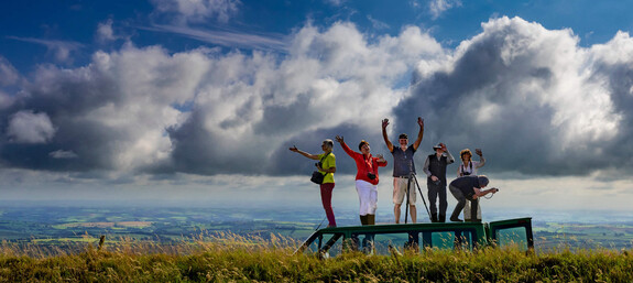 Fotogruppe steht auf Dach des Buses in grüner Exmoor-Landschaft und winkt begeistert Richtung Kamera. Weite und blauer Himmel mit Wolken.