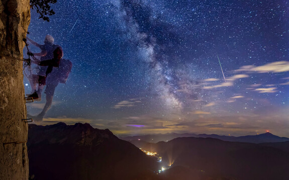 Hartmut Pönitz klettert nachts am Felsen der Steinplatte übern Klettersteig. Am Himmel die Milchstraße mit Sternschnuppen, unten die Lichter von Waidring.